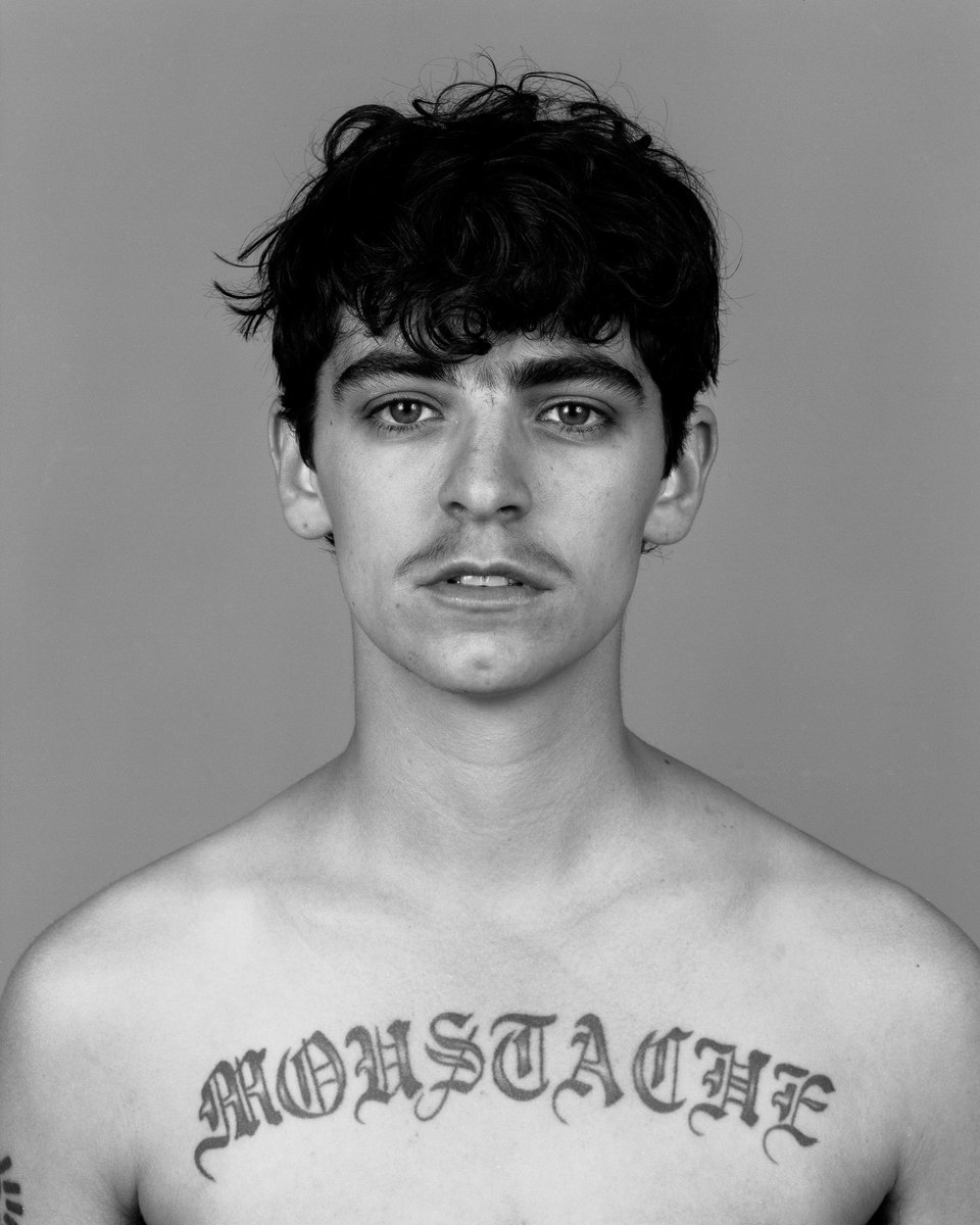 Monochrome portrait of a young adult with dark hair and a mustache, featuring a chest tattoo against a plain grey backdrop.