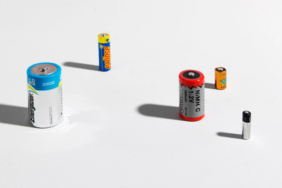 Assorted batteries including D, AA, AAA and C sizes are arranged on a white surface, casting shadows and highlighting various brands.