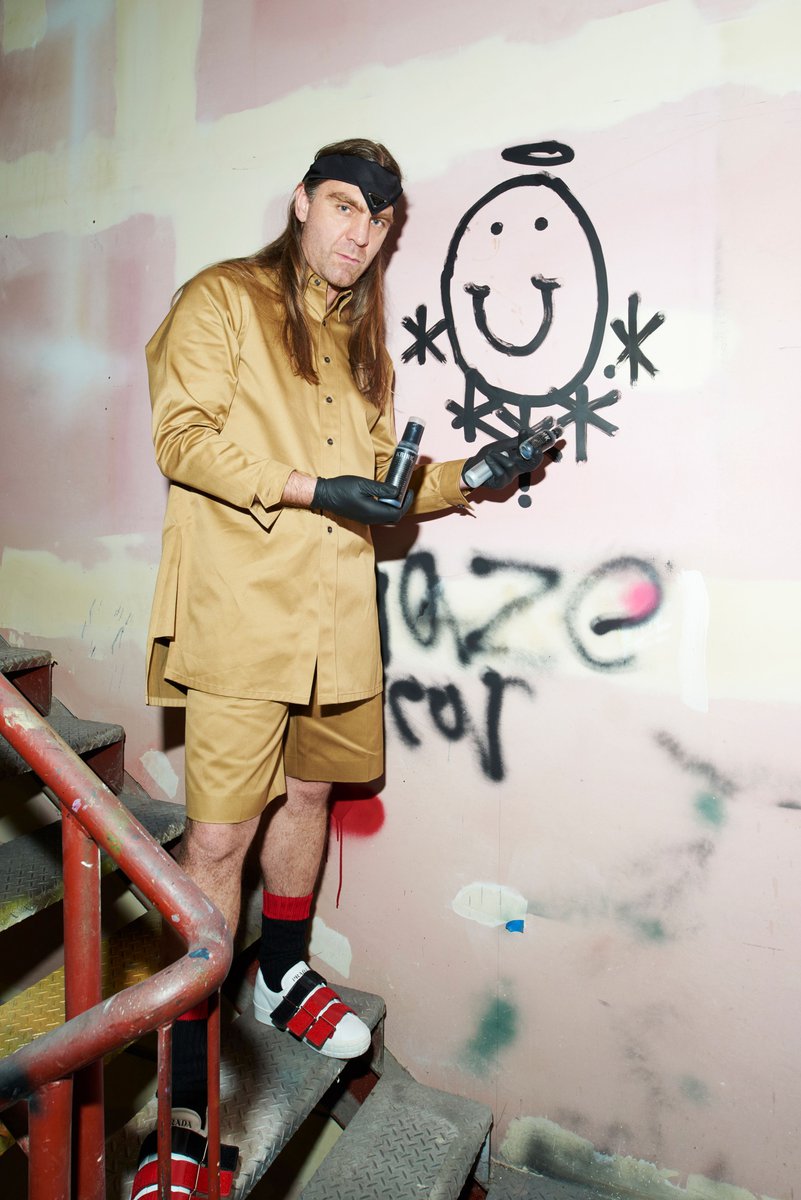 Man in tan jacket and shorts poses on stairs by graffiti wall holding spray cans, wearing black gloves, red and black socks.