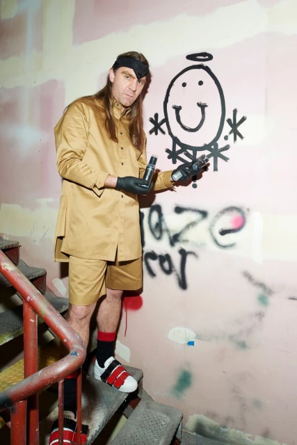 Man in tan jacket and shorts poses on stairs by graffiti wall holding spray cans, wearing black gloves, red and black socks.