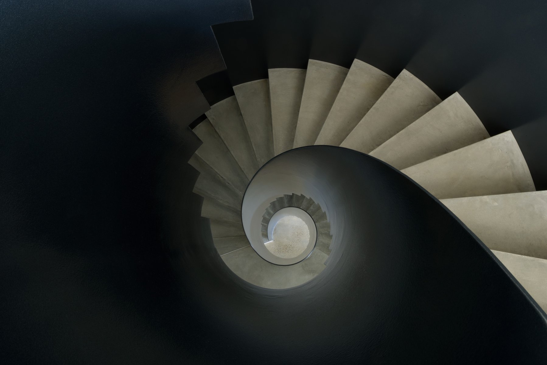Looking down at a spiral staircase with concrete steps and a dark gray bannister winding down to the center of the frame.