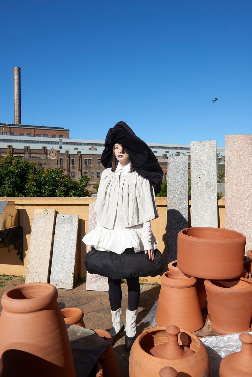 Fashionable young woman in a unique layered outfit, posing outdoors among terracotta pots and stone slabs, with a clear blue sky.