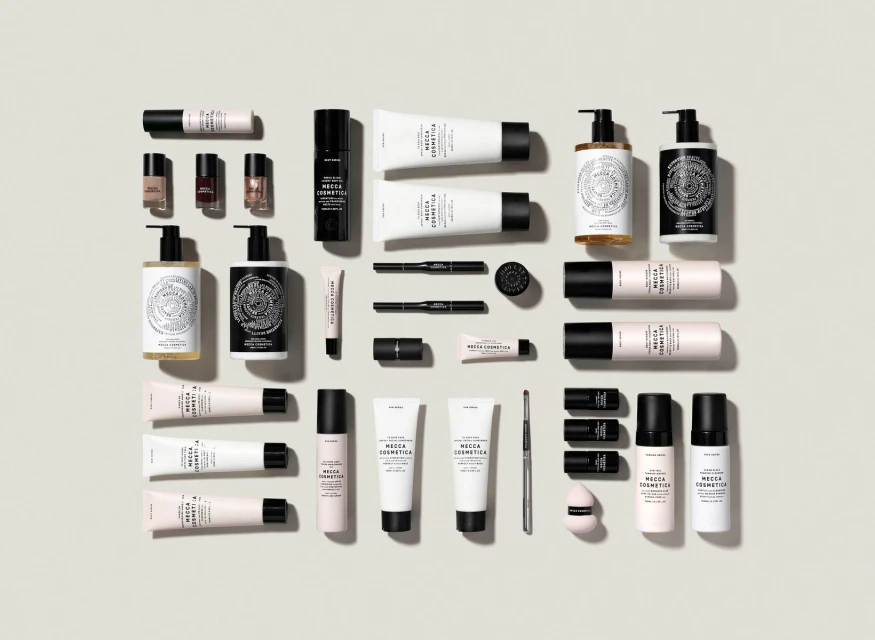 Flat lay of Mecca Cosmetica skincare, makeup, and beauty products in white, black, gold, and pink against a neutral backdrop.