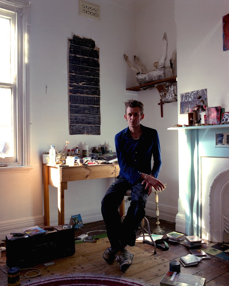 A man sits on a stool in a cluttered art studio with a large window, wooden desk covered with supplies, and art pieces on the walls.