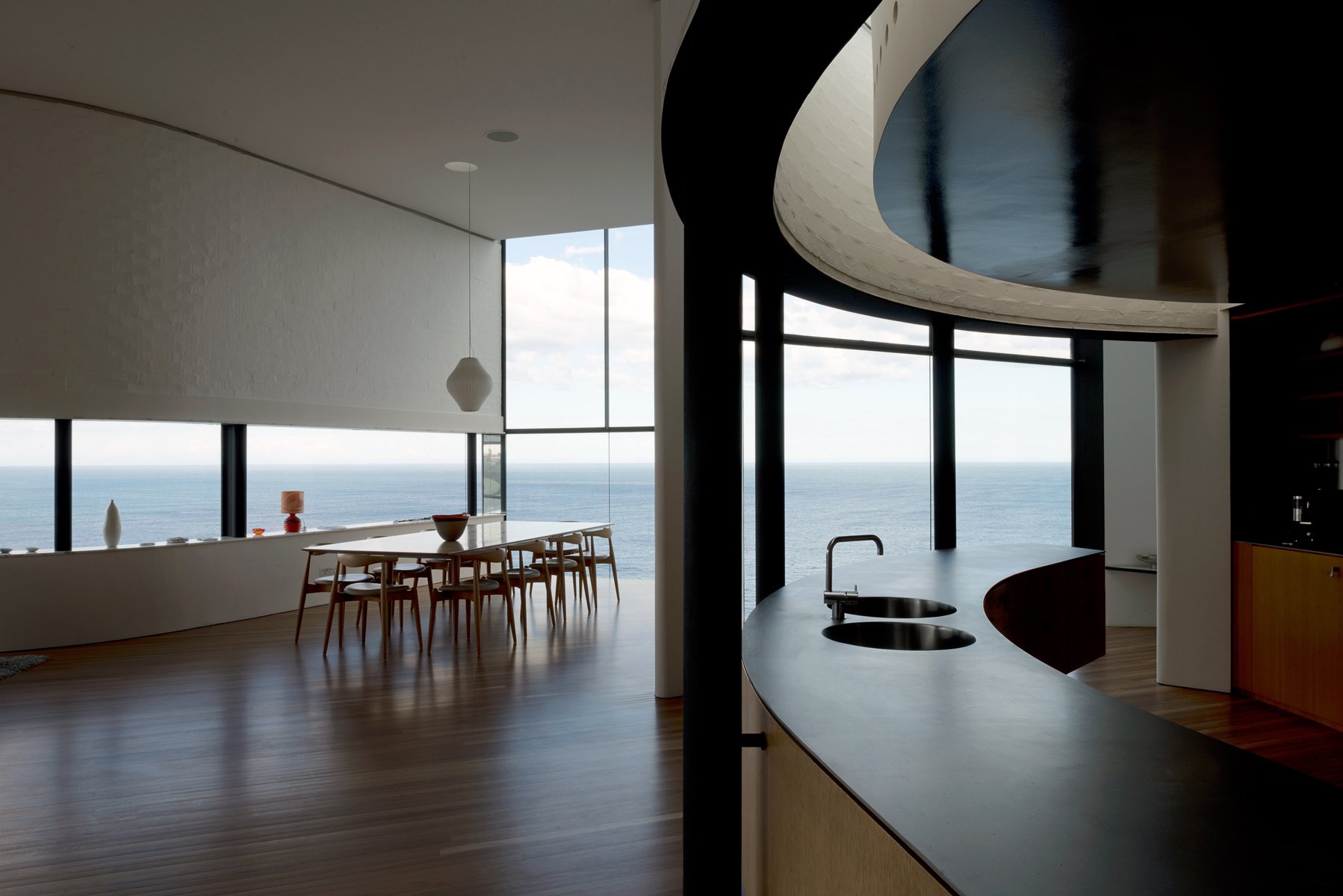 Modern home interior showcasing a dining table set near large windows with ocean views and a kitchen island with sinks.