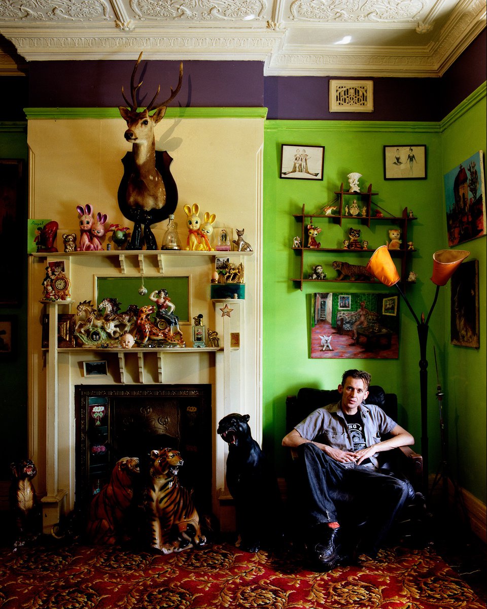 Interior shot of a man in a green and purple room seated in a black chair, flanked by animal statues, with shelves and a fireplace.