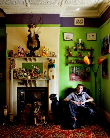 Interior shot of a man in a green and purple room seated in a black chair, flanked by animal statues, with shelves and a fireplace.