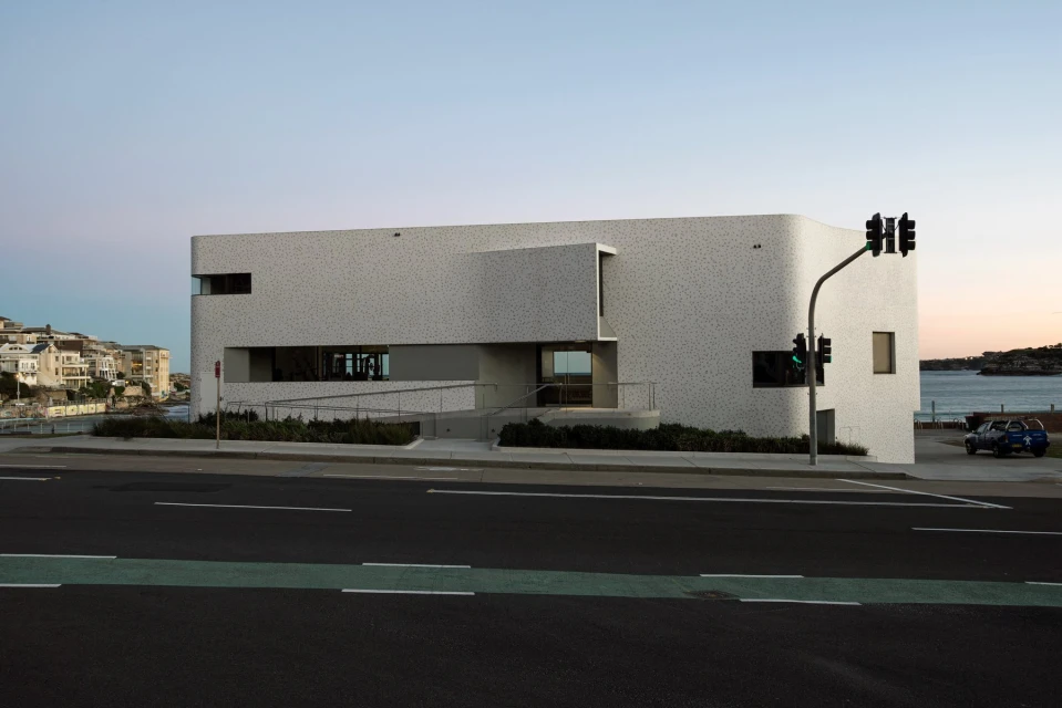 Exterior view of a modern, white building with geometric shapes and flat roof, set against a backdrop of ocean and sky at dusk.