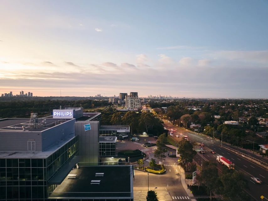 High-angle view of a modern building complex with 'Philips' signage on the roof, set against an urban skyline and a sunset sky.