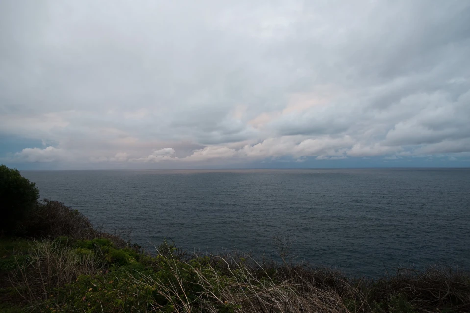 Cloudy seascape view of a dark ocean under a gray sky, with a grassy, elevated coastline in the foreground.