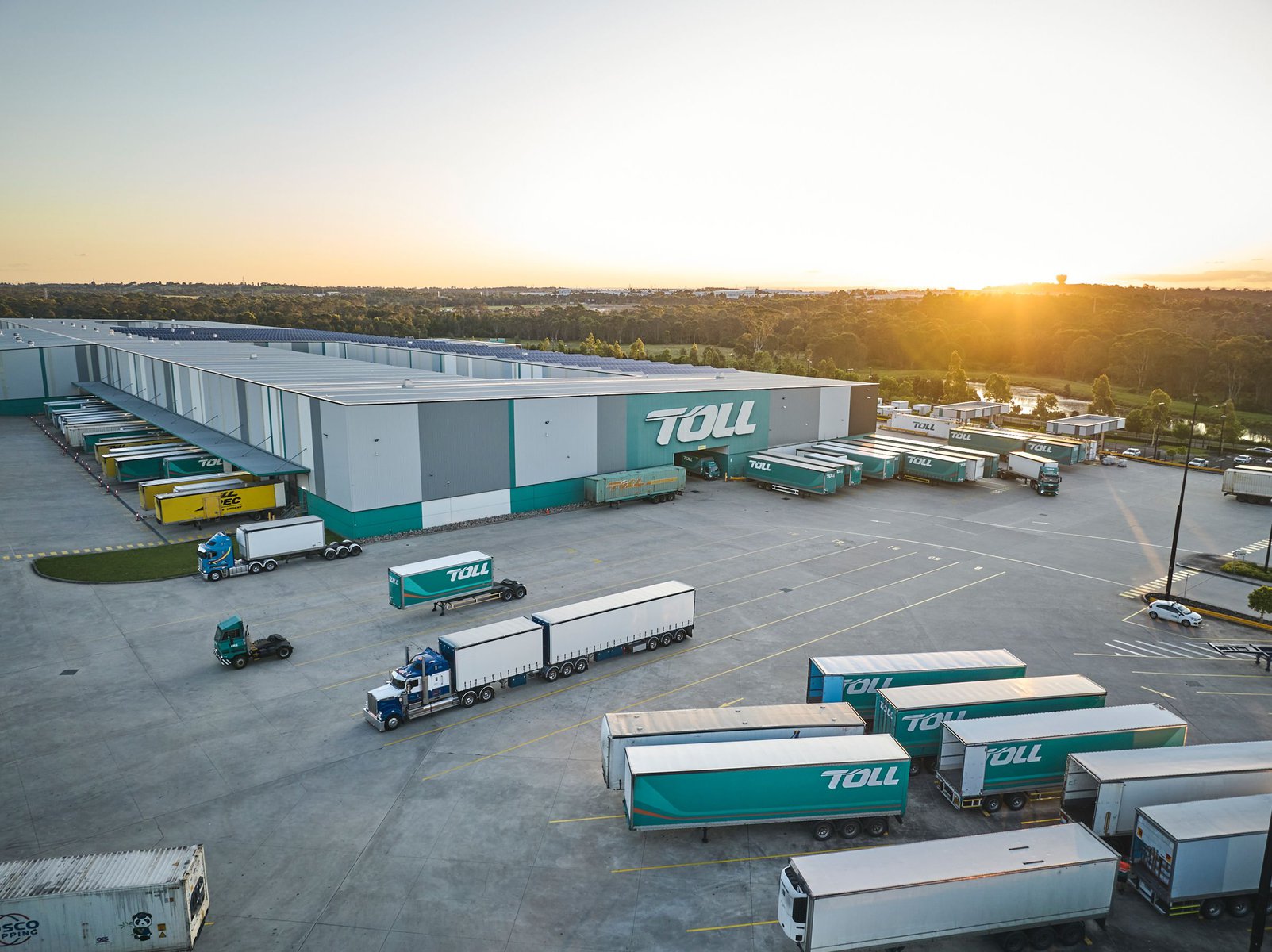 Aerial view of a logistics warehouse with numerous semi-trucks and trailers parked in the loading area, featuring the TOLL brand logo.