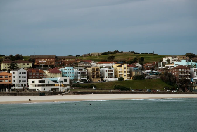 Coastal view of Bondi Beach in Australia, featuring buildings, the sandy shoreline, ocean waves, and a cloudy sky, showcasing a vibrant cityscape.