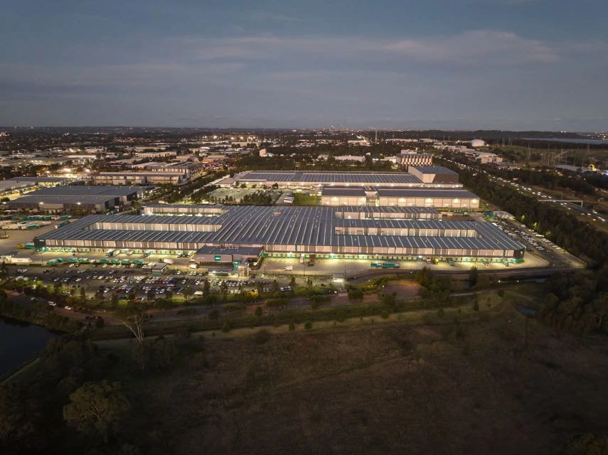 Aerial view captures a sprawling industrial complex at dusk, featuring large buildings with rooftop solar panels, surrounding greenery, and city lights.