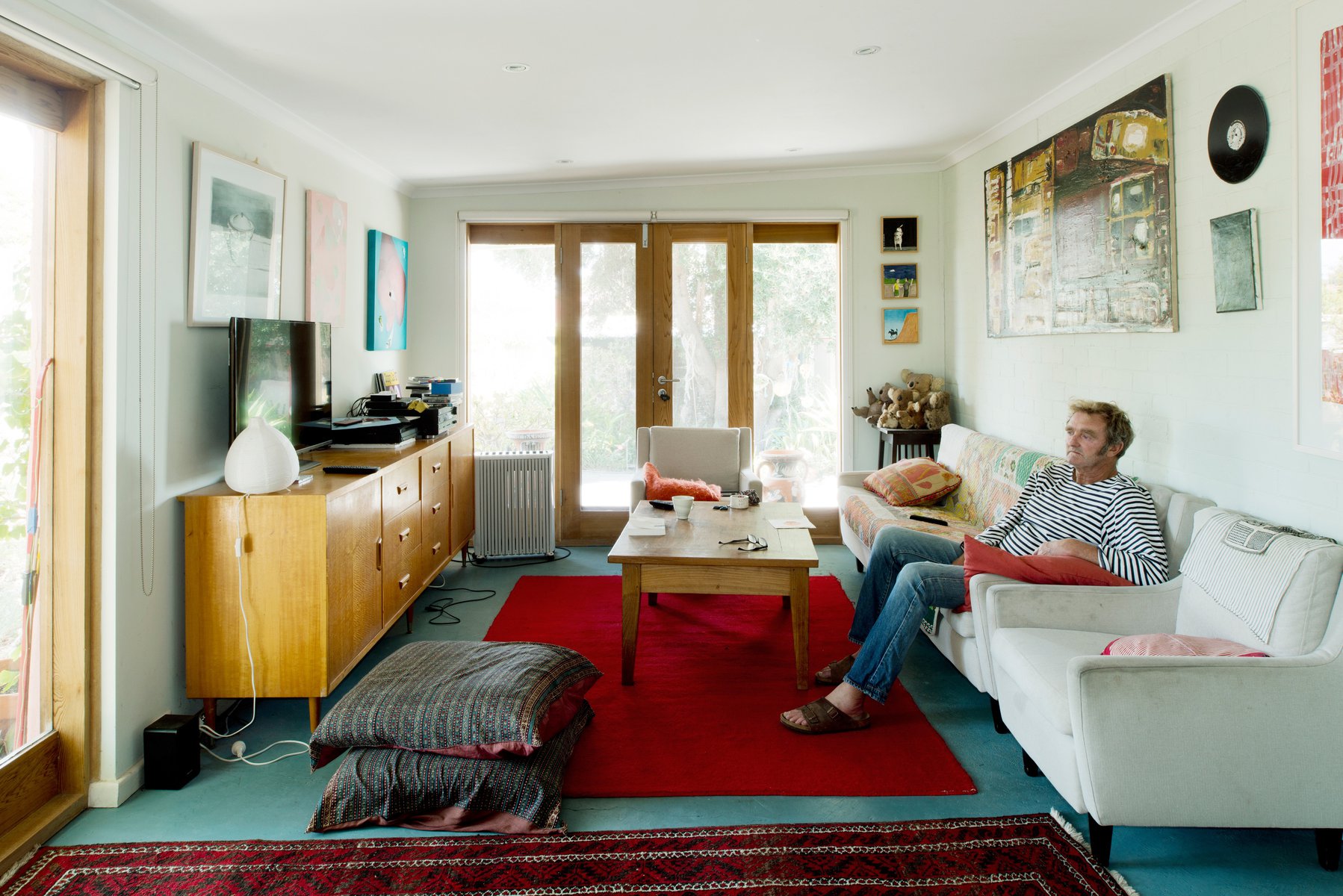 A man lounges on a white couch in a sunlit living room with light blue walls, a red rug, and artwork on the walls.