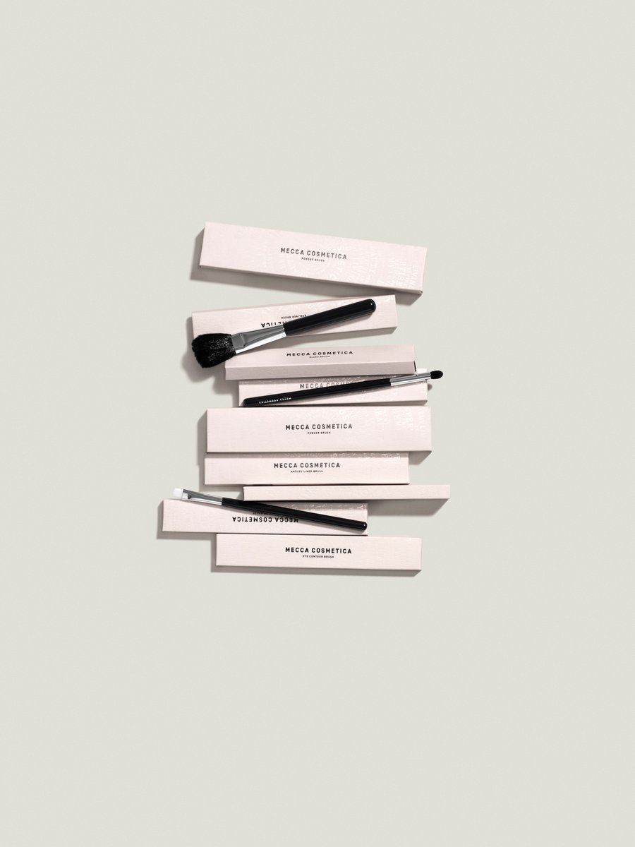 Overhead shot of several makeup brushes with black handles stacked on top of light pink boxes, on a light grey background.