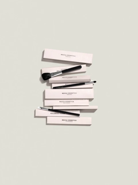 Overhead shot of several makeup brushes with black handles stacked on top of light pink boxes, on a light grey background.