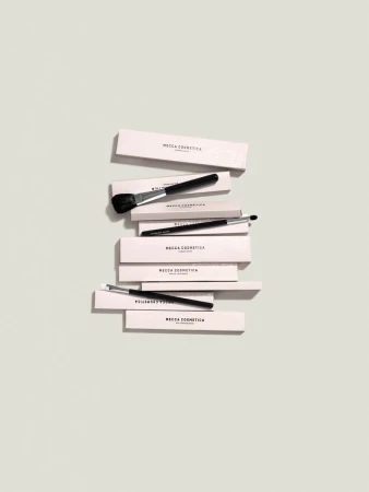 Overhead shot of several makeup brushes with black handles stacked on top of light pink boxes, on a light grey background.