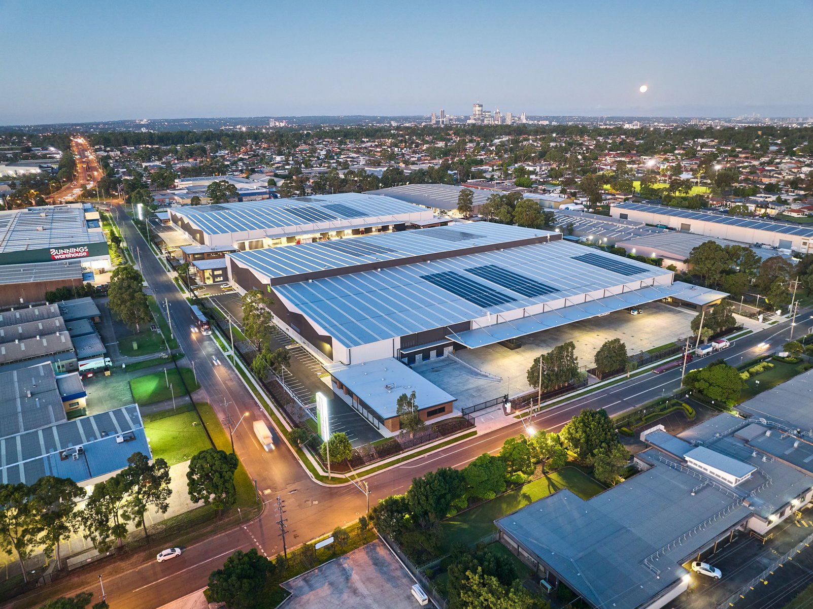Aerial view of modern industrial buildings with solar panels on the roof, surrounded by green trees and roads under a clear sky at dusk.