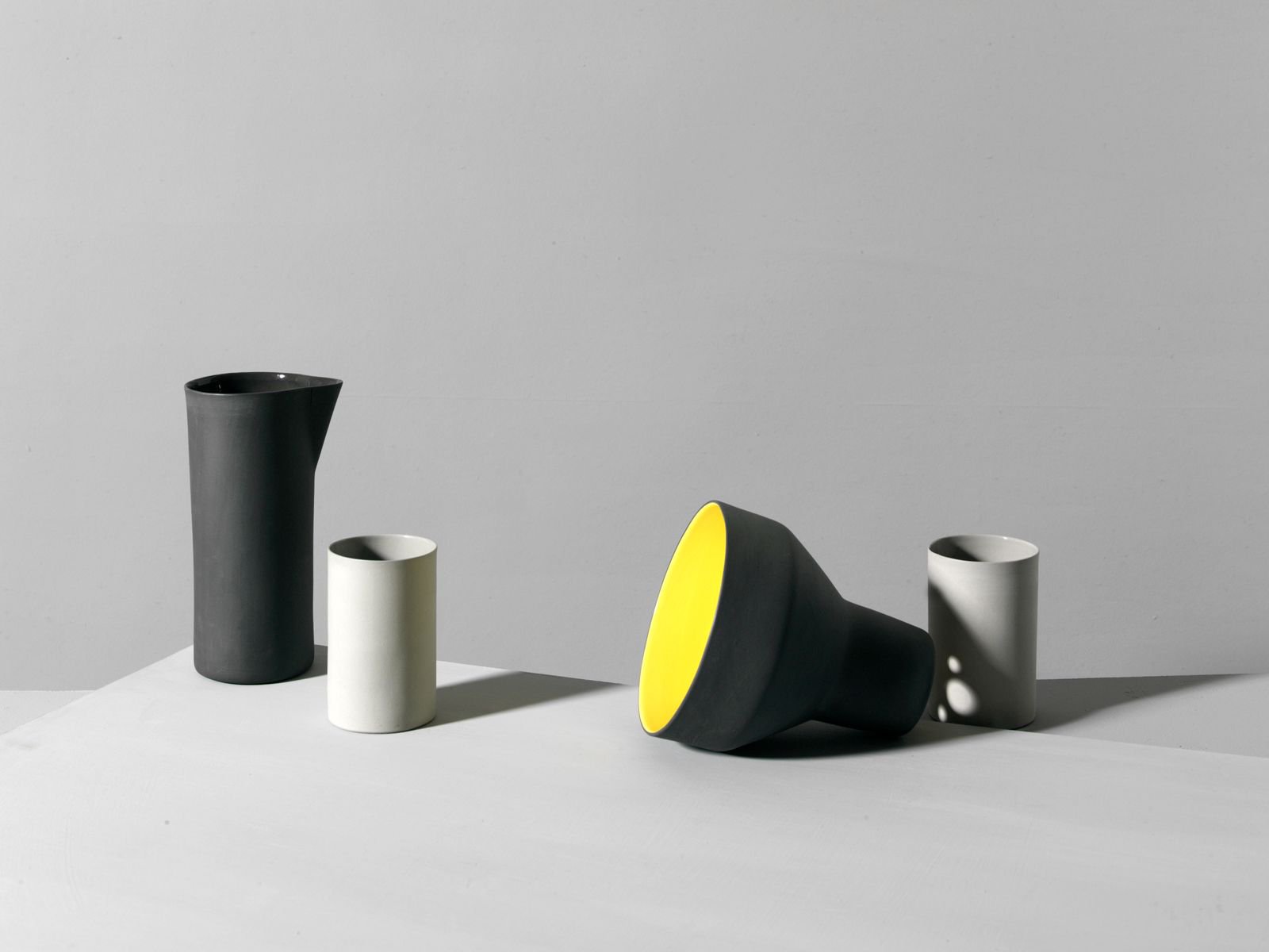 Still life features matte black and white ceramic jugs, arranged on a bright white surface against a light gray studio background.
