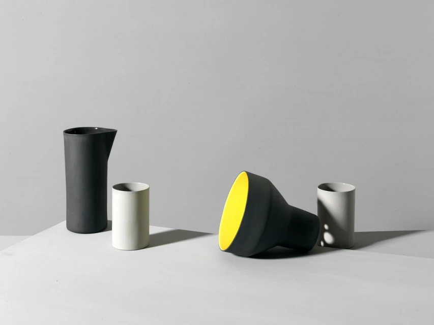 Still life features matte black and white ceramic jugs, arranged on a bright white surface against a light gray studio background.