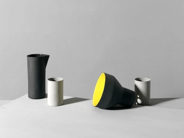 Still life features matte black and white ceramic jugs, arranged on a bright white surface against a light gray studio background.