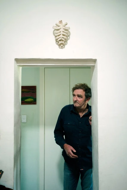 Man stands in doorway of white-walled room with painting; wall decoration resembling bone structure is hung near the door.