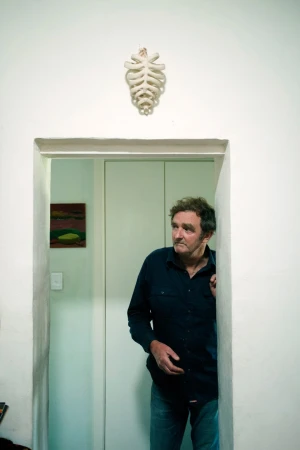 Man stands in doorway of white-walled room with painting; wall decoration resembling bone structure is hung near the door.