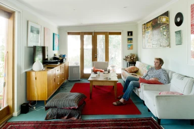 Man seated on a white sofa in a brightly lit living room with wooden furniture and a vibrant red area rug.