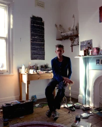 Man seated on stool in a cluttered room with a desk, fireplace, and window lighting by photographer Andrew Cowen