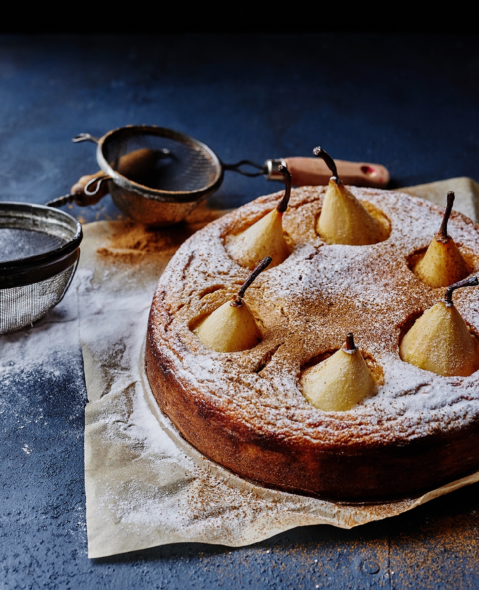Overhead view of a pear cake dusted with powdered sugar, featuring whole pears baked inside, presented with a sieve on baking paper.