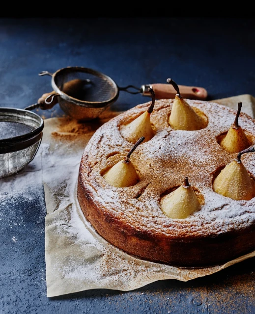 Overhead view of a pear cake dusted with powdered sugar, featuring whole pears baked inside, presented with a sieve on baking paper.