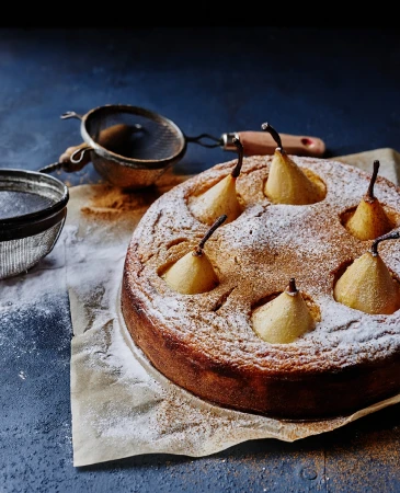 Overhead view of a pear cake dusted with powdered sugar, featuring whole pears baked inside, presented with a sieve on baking paper.