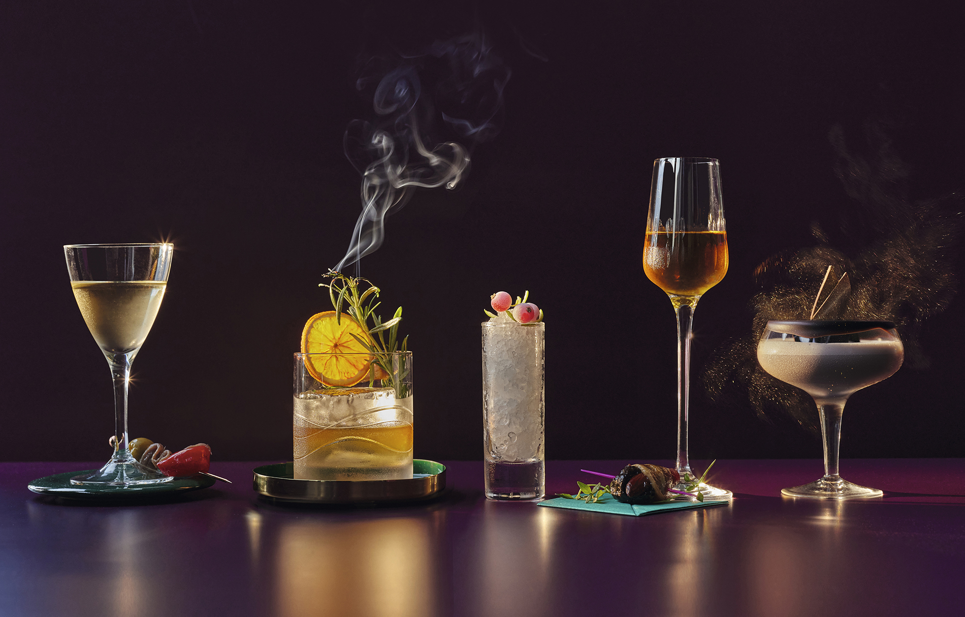 Five cocktails in stemmed glasses, featuring citrus, herbs, berries, and a smoky vapor, arranged on a glossy purple surface.