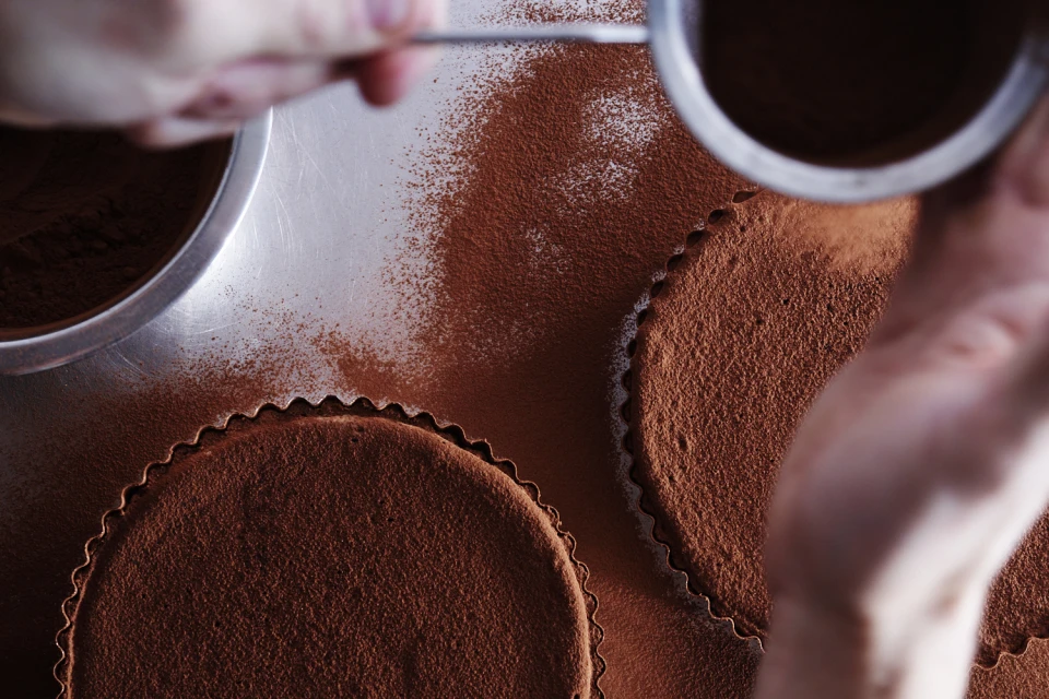 Overhead shot of tarts being dusted with cocoa powder, showcasing the baking process and rich color of the dessert ingredient.