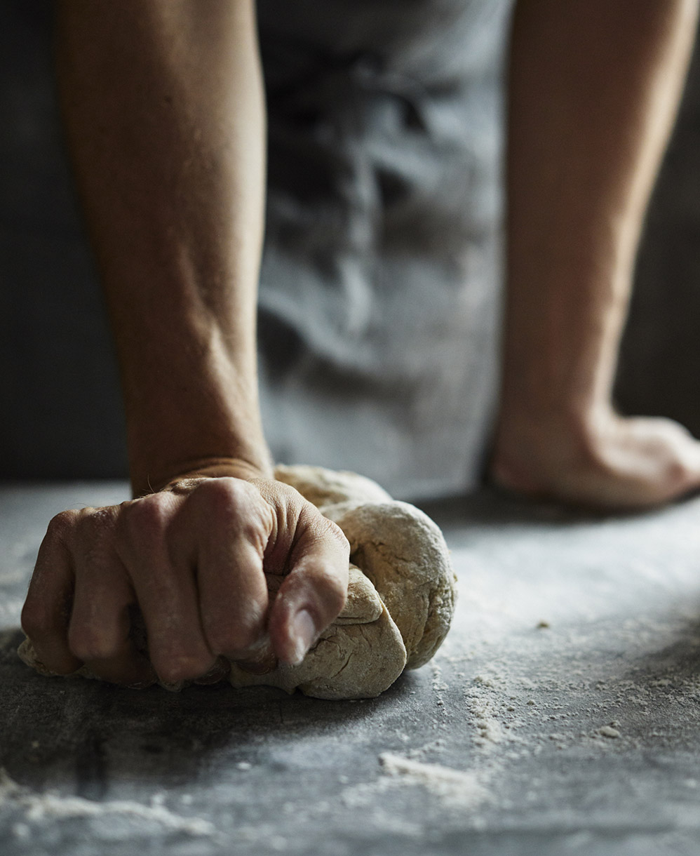 Person kneading dough on a flour-dusted counter. Close-up shot of hands working the dough, set against a muted, dark gray background.