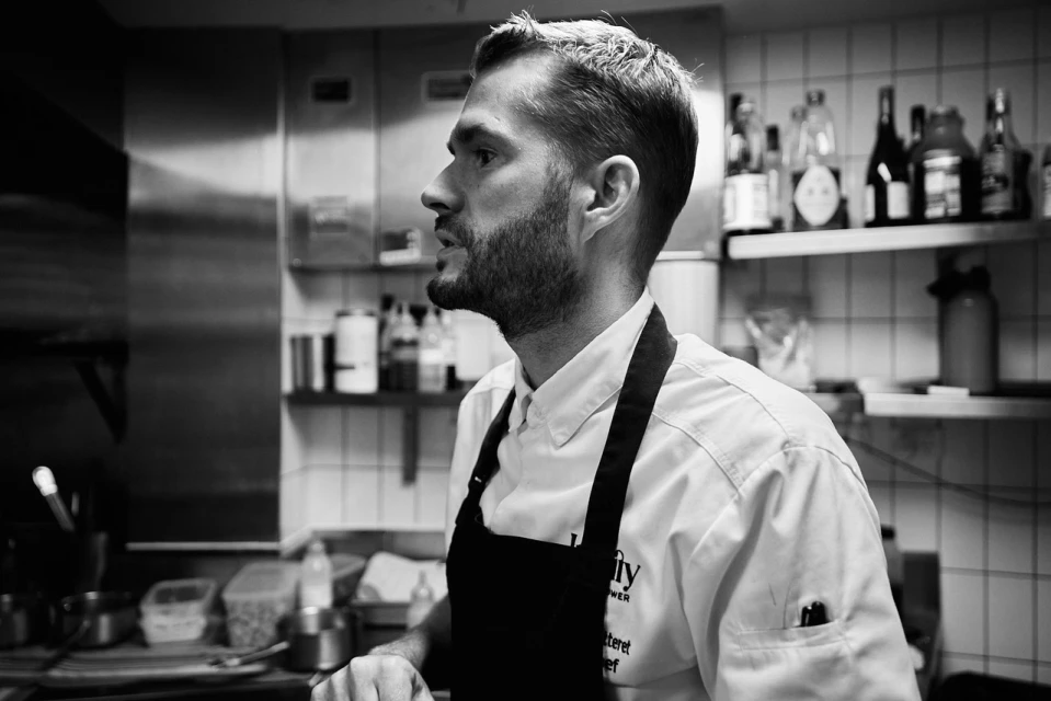 Profile shot of a bearded adult male chef wearing a black apron in a commercial kitchen with stainless steel equipment and tiled walls.