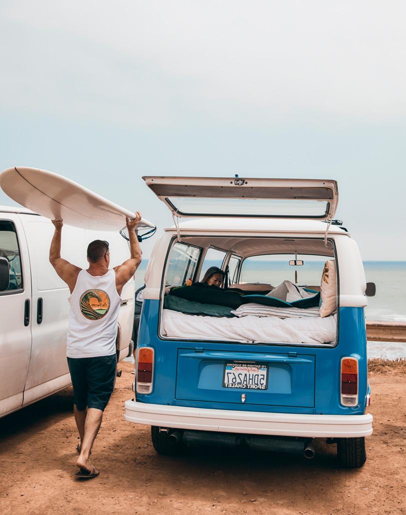 Man lifts a surfboard next to a VW van at the beach, with another person visible inside the open back hatch and blue ocean visible.