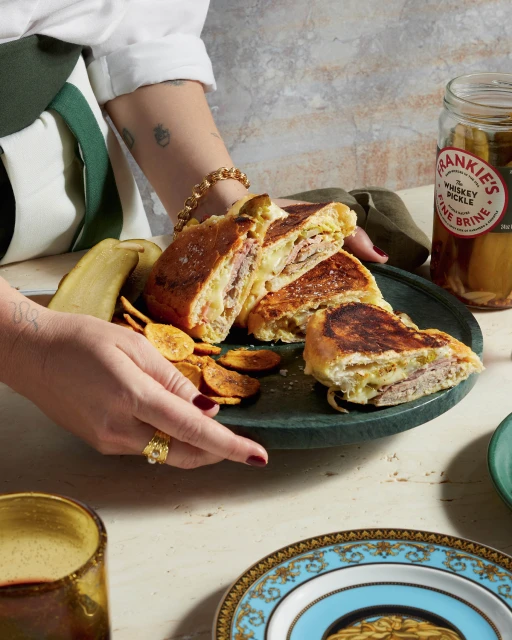 Hands holding a plate of sliced Cubano sandwiches, with pickles and chips, arranged on decorative plates on a marble surface.