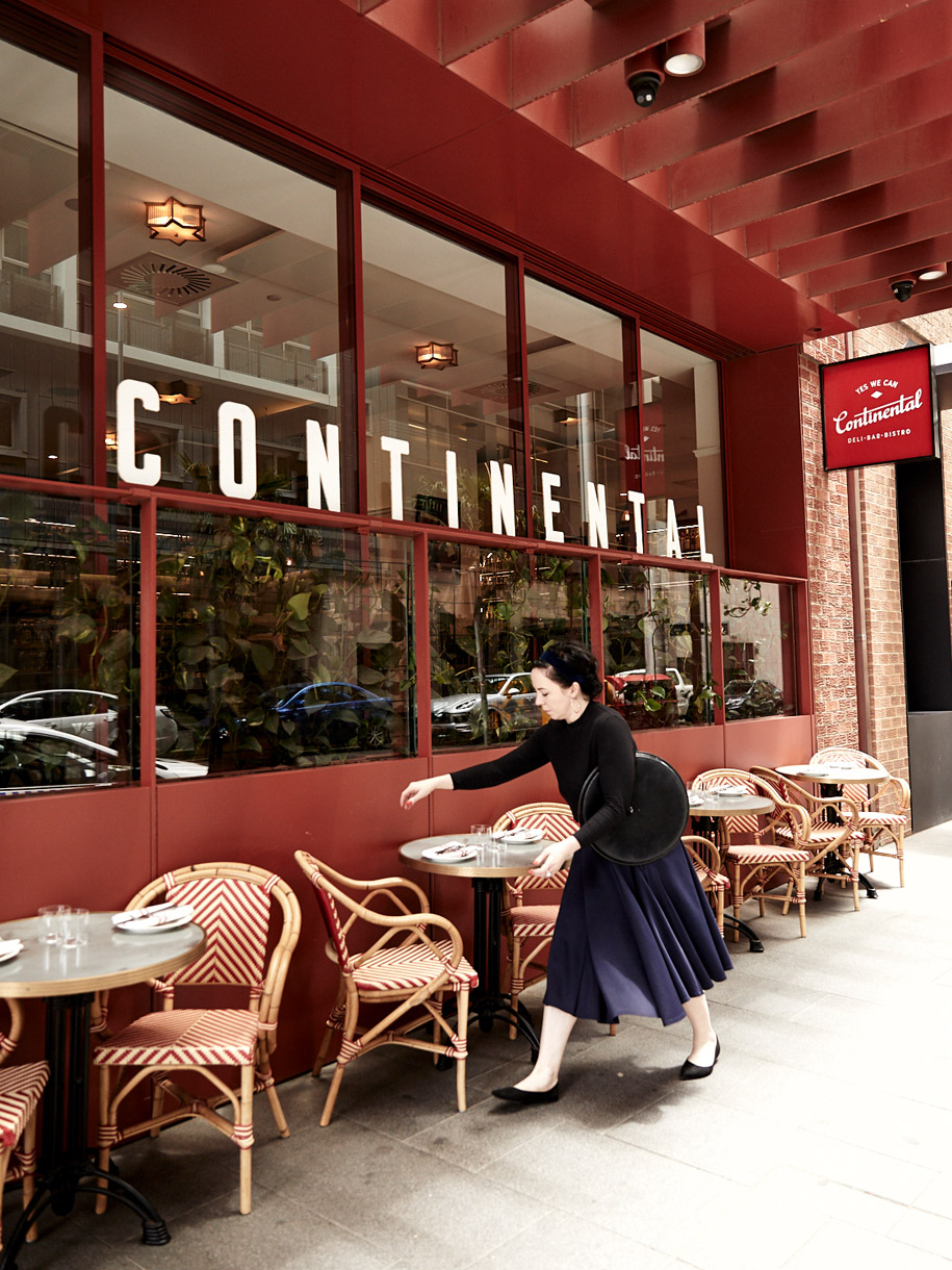 Adult female in a dark top and navy skirt sets a table outside the Continental bistro on a partly cloudy day, with cane chairs.