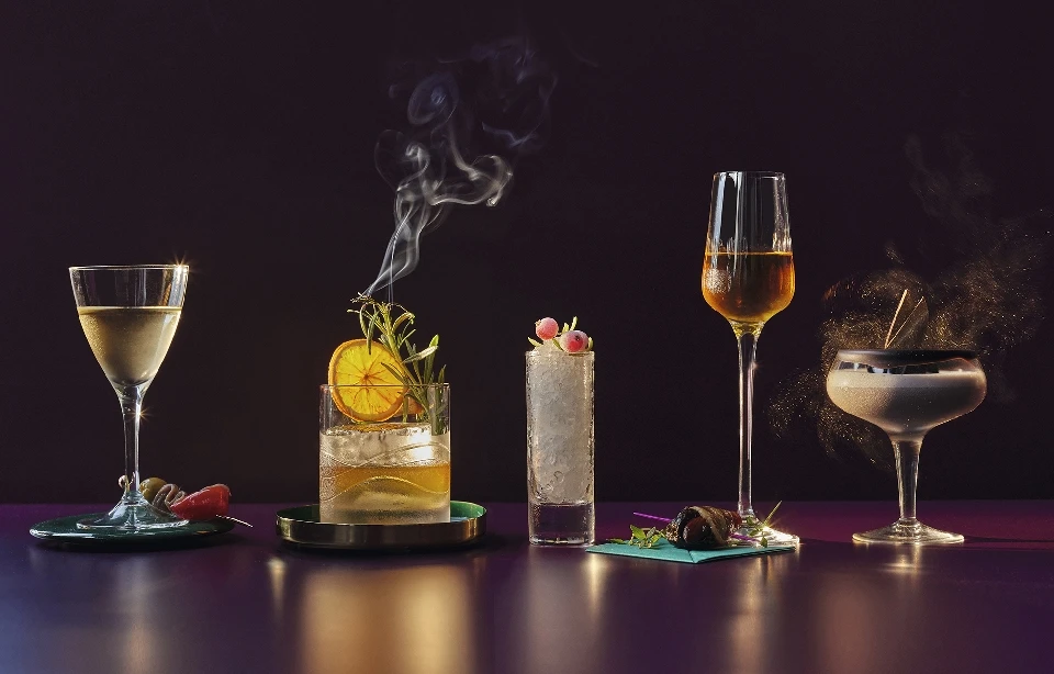Five cocktails of varying styles are lined up in a studio setting, on a reflective purple surface against a dark background.