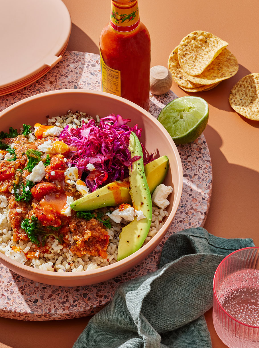 Overhead shot of a vibrant grain bowl with avocado, red cabbage, kale, tomatoes, and crumbled cheese on a colorful background.