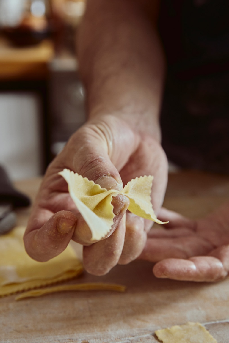 Close-up of handmade farfalle pasta held in a person's hand with more fresh pasta and a wooden cutting board visible in the background.