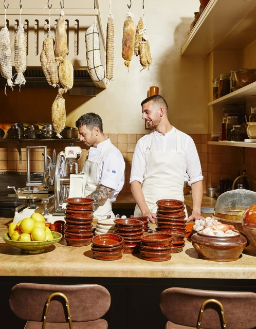 Two chefs in a restaurant kitchen prepare food; terracotta bowls are stacked on a countertop with cured meats hanging above.