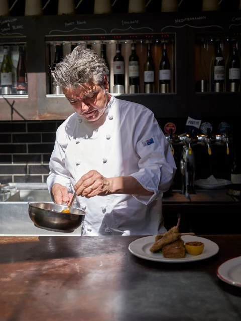 Chef in a white coat prepares food in a restaurant, using a spoon in a steel bowl, with plates and wine bottles in the background.
