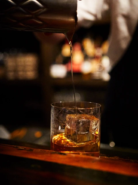Bartender pours amber liquid from a metal shaker into a clear glass with a large ice cube, set on a wood bar top.