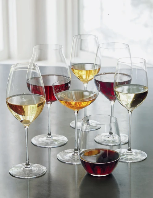 Wine glasses filled with red, white, and rose wine stand on a table in front of a window with sheer white curtains.