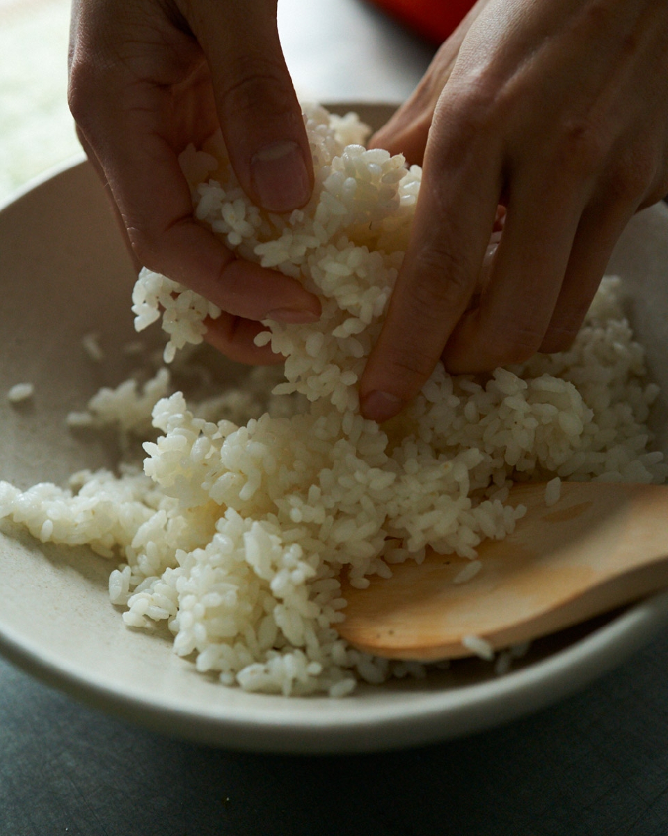 Close-up of hands mixing white rice in a bowl with a wooden spoon; food preparation in indoor kitchen setting.