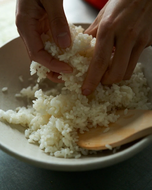Close-up of hands mixing white rice in a bowl with a wooden spoon; food preparation in indoor kitchen setting.