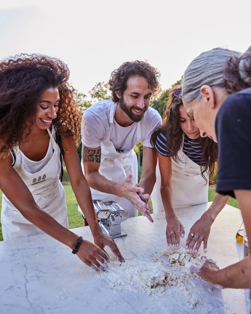A group of four people, including one adult male and three adult females, gathered outdoors around a table making pasta, smiling and engaging.
