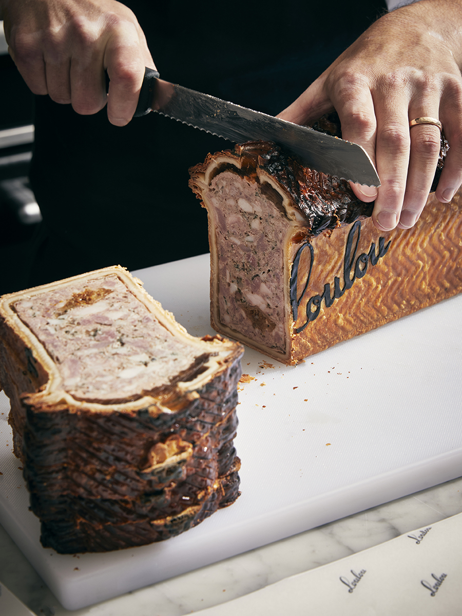 Chef slices a meat pâté en croûte with a serrated knife on a white cutting board, featuring the word "Loulou" on the crust.
